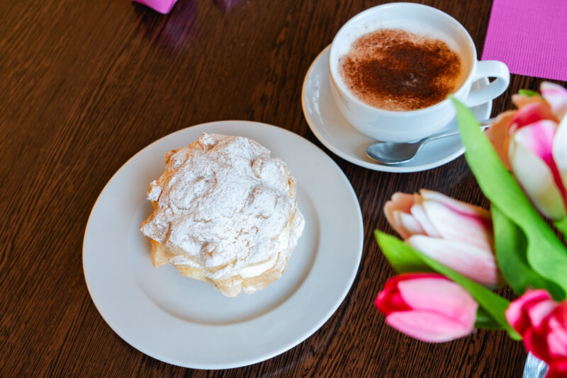 Draufsicht auf einen Windbeutel mit Puderzucker auf weißem Teller, daneben eine Tasse Cappuccino mit Kakaostreuseln und einem Strauß pinker Tulpen auf einem dunklen Holztisch.