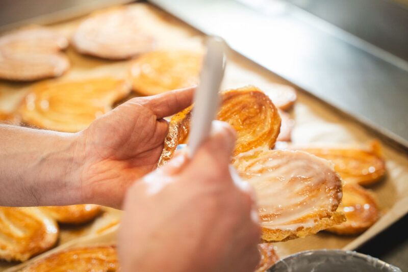 Nahaufnahme zweier Hände beim Glasieren von frisch gebackenen Palmiers (Schweineohren) mit einem Zuckerguss-Pinsel auf einem Backblech mit Backpapier