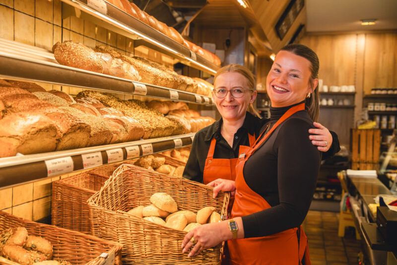 Zwei Mitarbeiterinnen der Bäckerei Dreimann in schwarzen Oberteilen und orangefarbenen Schürzen posieren lächelnd hinter dem Tresen, vor einem gefüllten Brötchenkorb und gut bestückten Brotregalen.